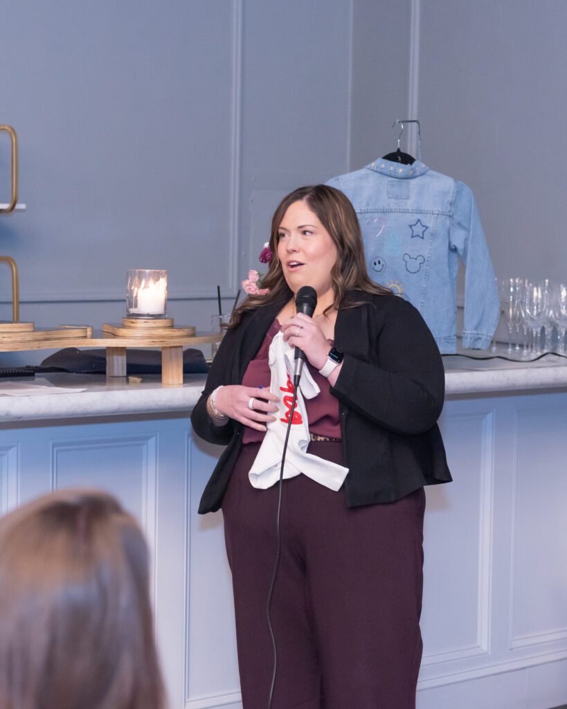 A woman is holding a mic and baby onesies while giving a speech at an event.