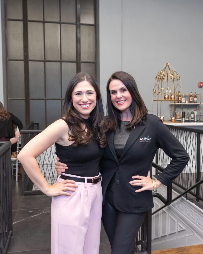 Two women standing with their one hand on their hips and smiling at an embroidering event.