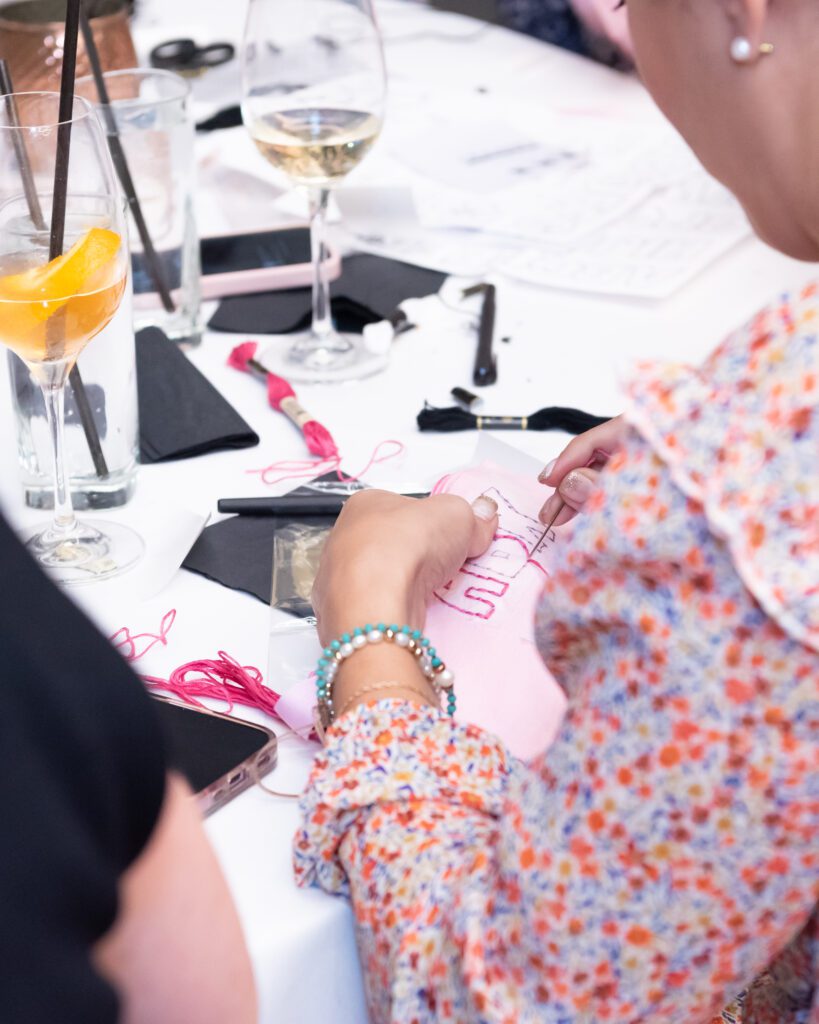 A woman wearing a floral top is sitting and hand embroidering a pouch.