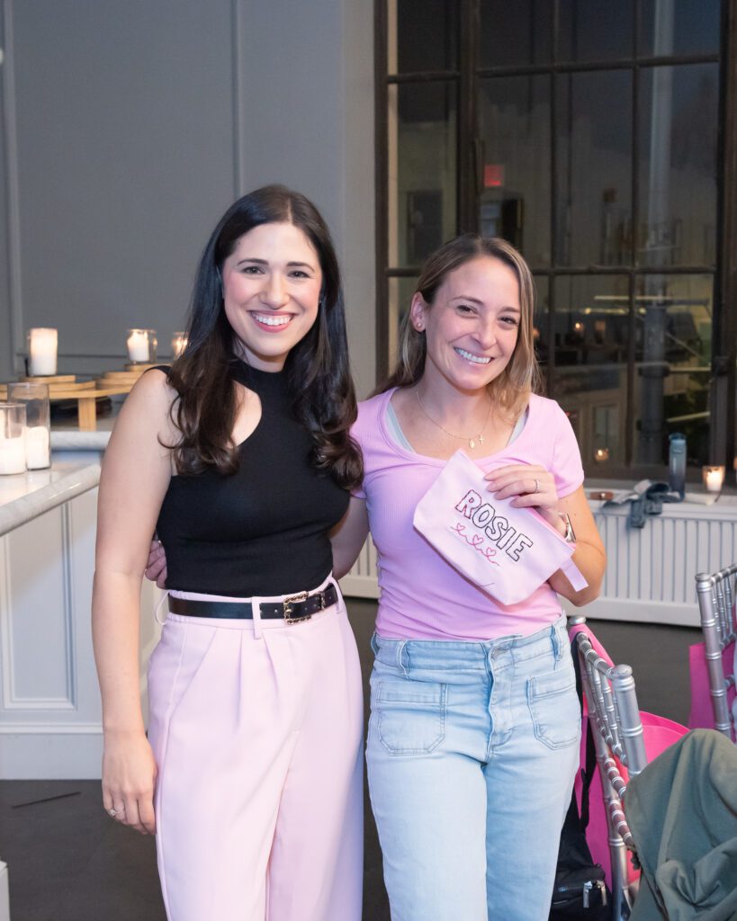 Two women standing and smiling and one on the right is holding a pouch that she just hand embroidered.