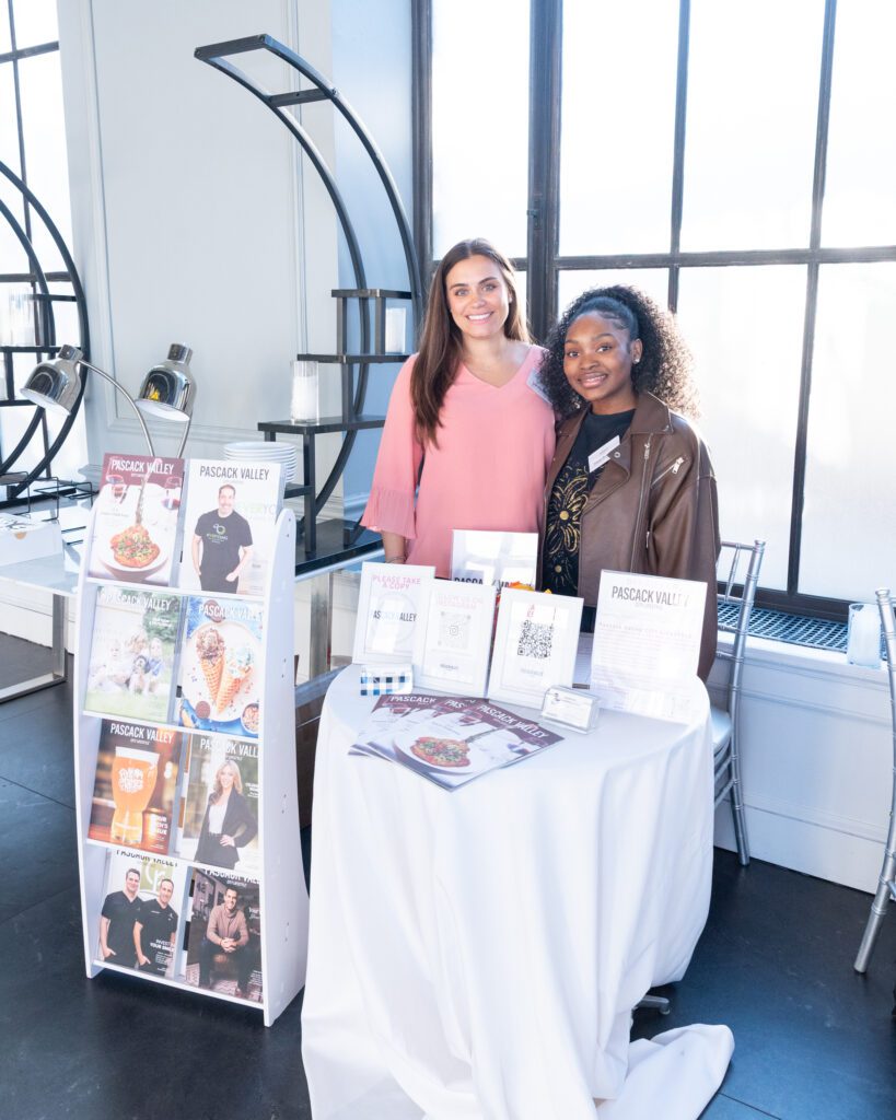 Two women from the Pascack Valley City Lifestyle magazine standing and posing at their vendor table.