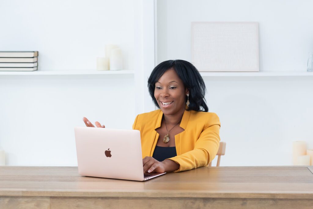 A female social entrepreneurship coach wearing a gold blazer and black dress sitting and talking to her client on her laptop at her office.