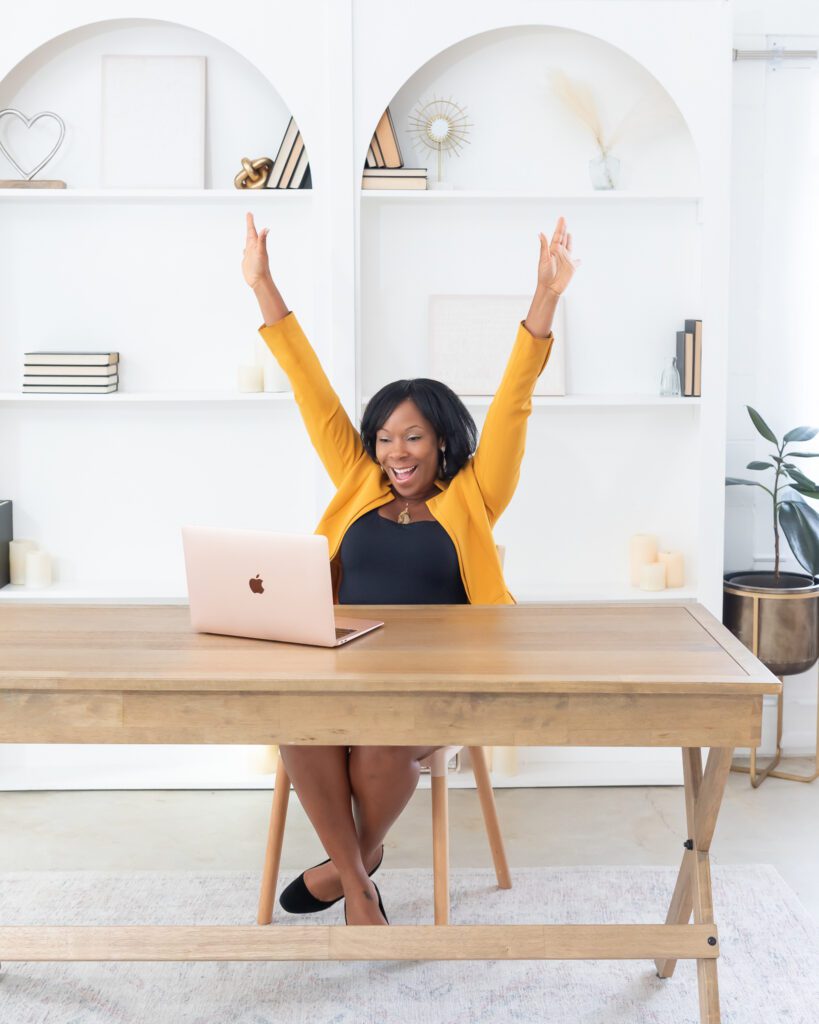A female social entrepreneurship coach wearing a gold blazer and black dress sitting in her office with her laptop, and cheering for her client on a video conference.