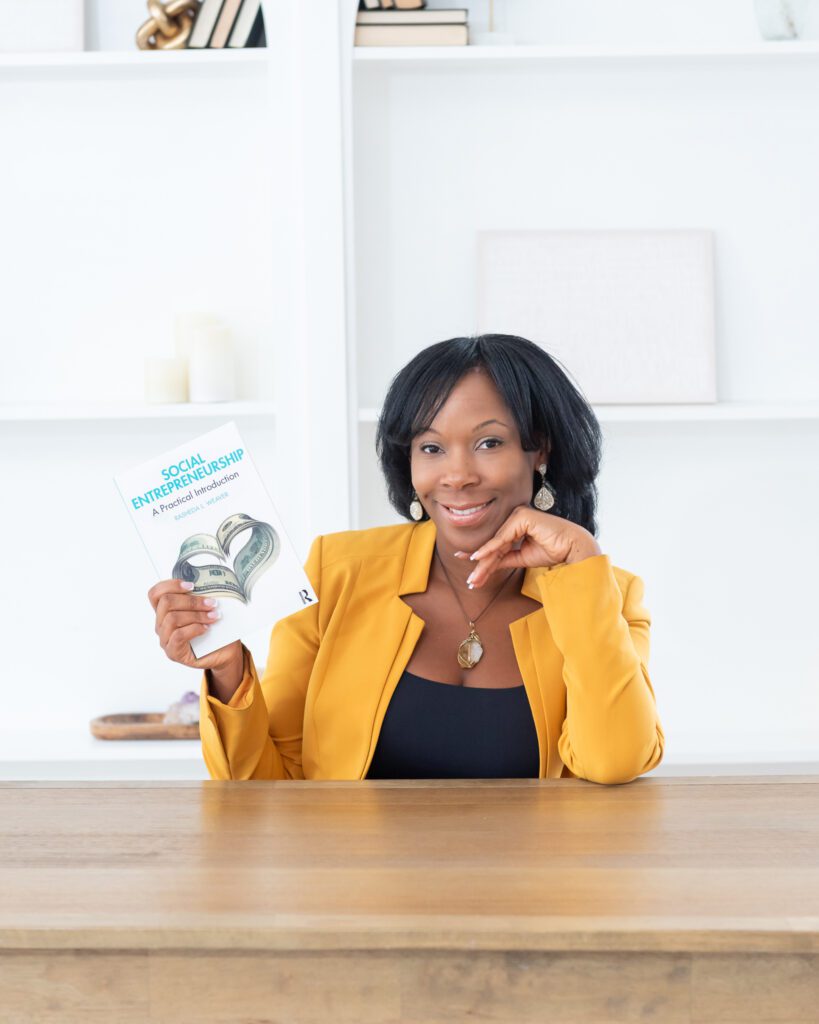 A female social entrepreneurship coach wearing a gold blazer and black dress sitting at her desk, smiling, and holding her book.