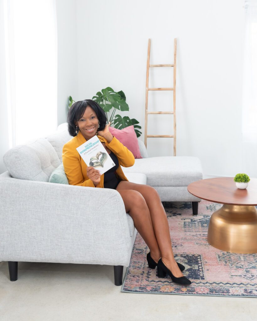 A female social entrepreneurship coach wearing a gold blazer and black dress sitting on a gray couch, smiling, and holding her book.