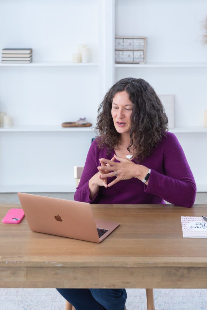 A female executive & leadership coach wearing a purple sweater talking on a video conference with her client in her office.