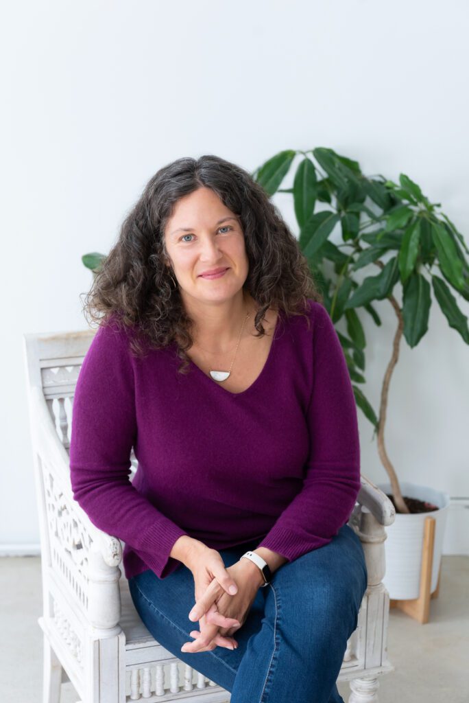 A female executive & leadership coach wearing a purple sweater sitting and smiling.