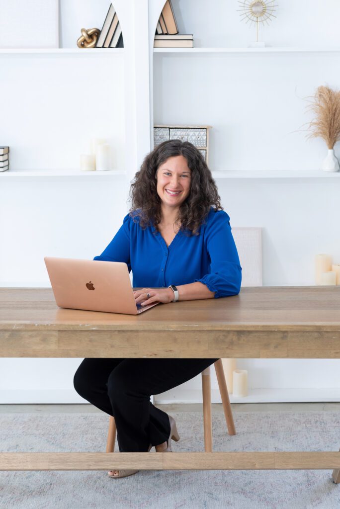 A female executive & leadership coach wearing a blue blouse working on her laptop and smiling in her office.