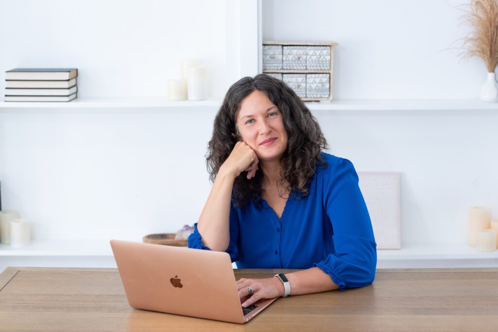 A female executive & leadership coach wearing a blue blouse working on her laptop and smiling in her office.