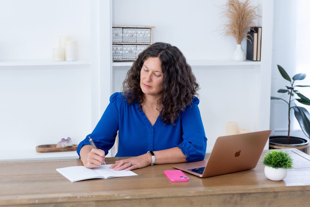 A female executive & leadership coach wearing a blue blouse writing in her notebook in her office.