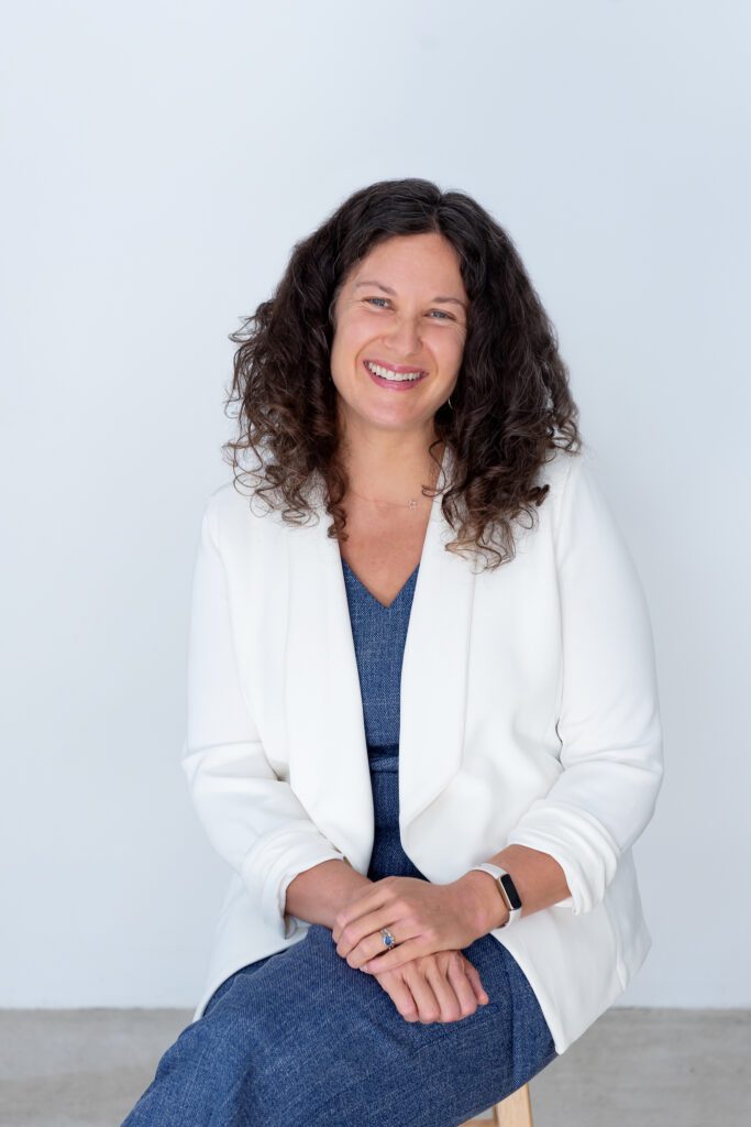 A female executive & leadership coach wearing a jumper suit and a white blazer sitting and smiling.