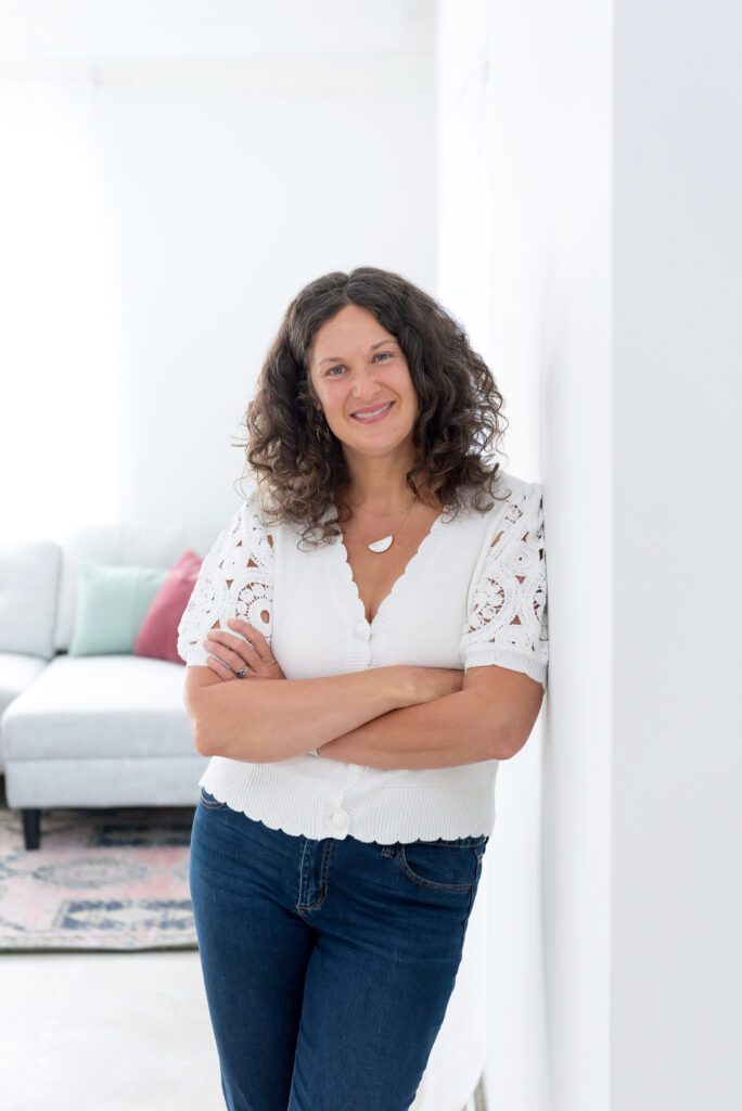 A female executive & leadership coach wearing a white top and jeans with her arm crossed and smiling in her living room.