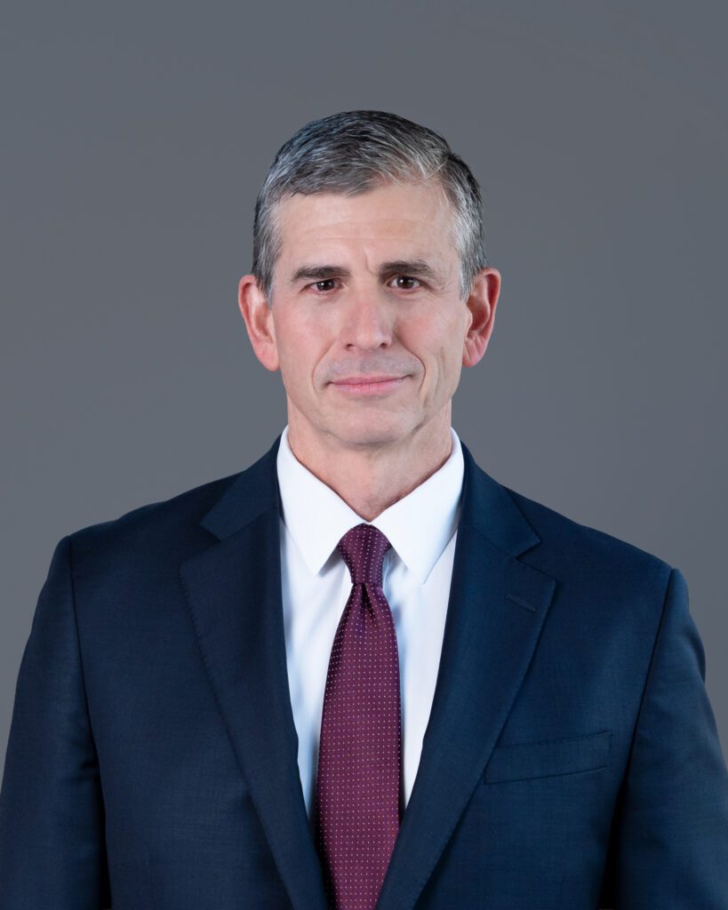 A male public speaker in a dark blue blazer and maroon color tie at his headshot session with a gray backdrop.