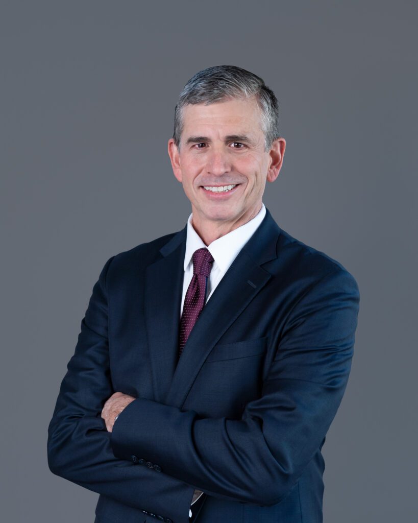 A male public speaker smiling with his arm crossed for his headshot session with a gray backdrop.