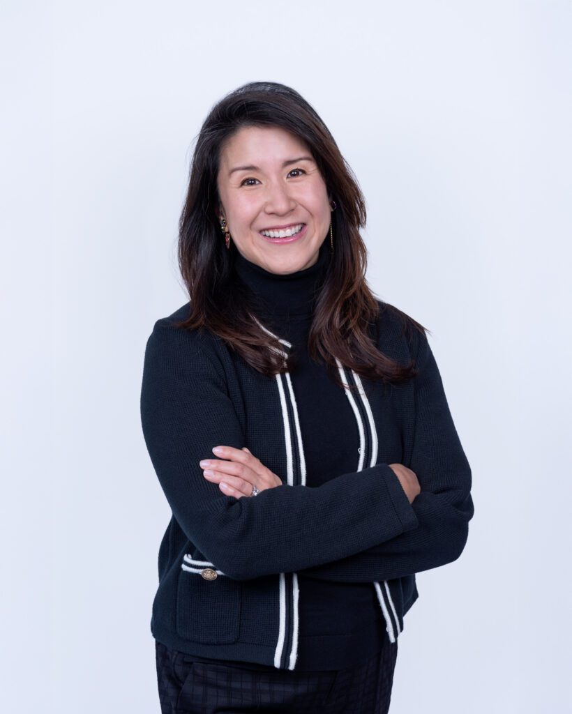 A female Asian executive coach & advisor smiling with her arm crossed for her headshot session with a white backdrop.