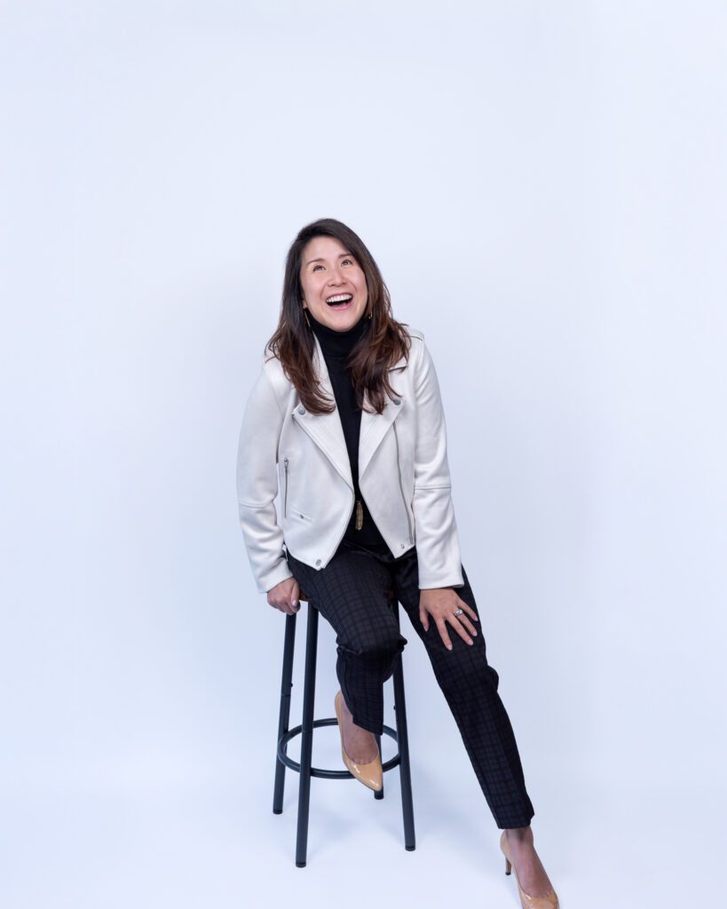 A female Asian executive coach & advisor laughing and sitting on a bar stool for her headshot session with a white backdrop.