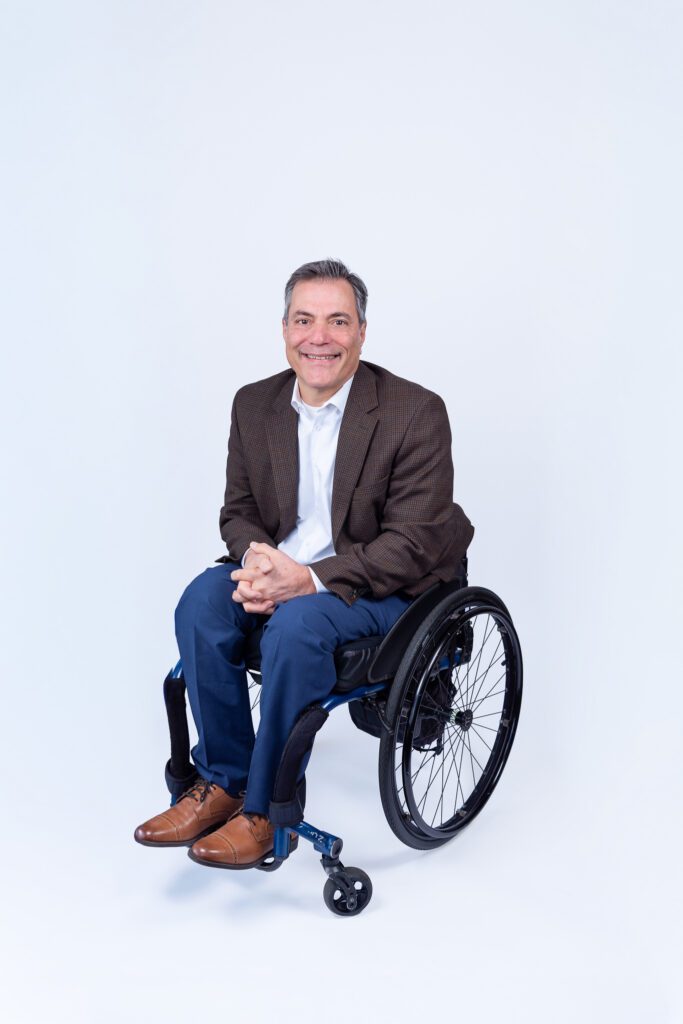 A male public speaker in a wheel chair smiling for his headshot session with a white backdrop.