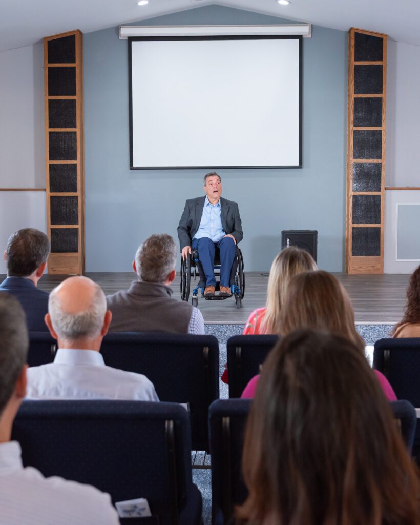 A male public speaker in a wheel chair talking on the stage with a live audience.