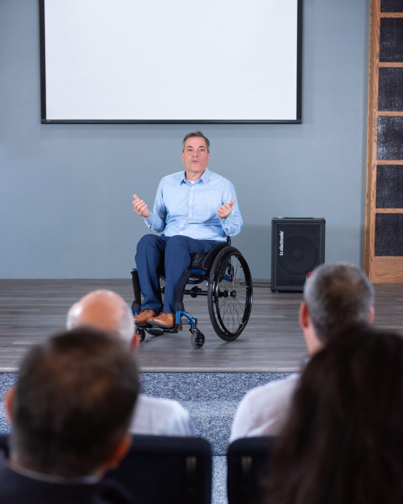 A male public speaker in a wheel chair talking on the stage with a live audience.