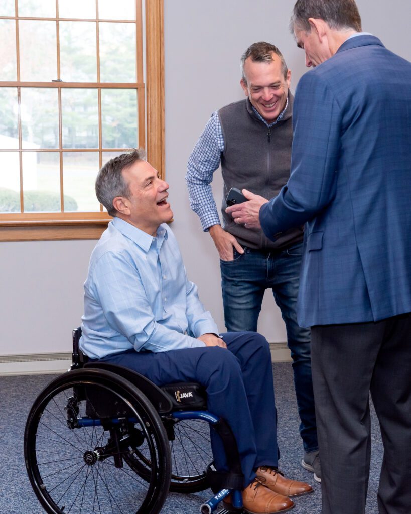 A male public speaker in a wheel chair talking and laughing with two male attendees at an event.