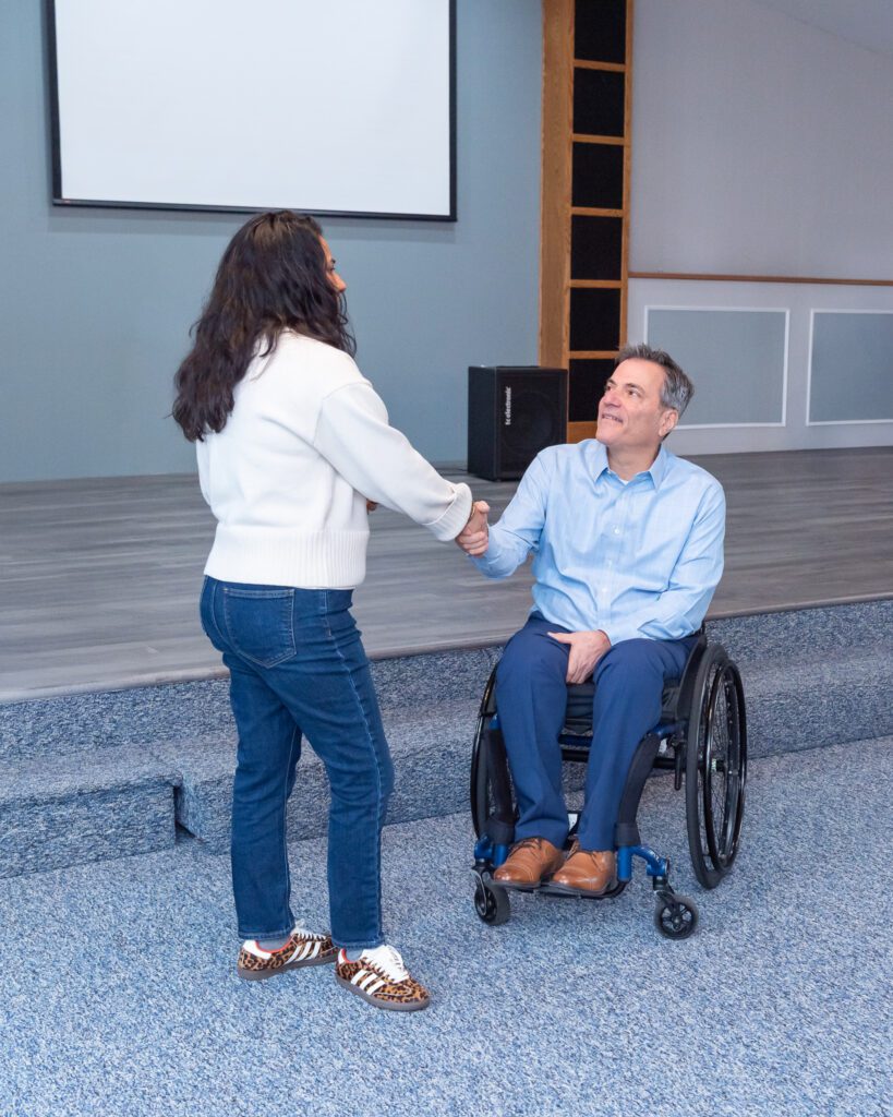 A male public speaker in a wheel chair shaking hand with a female attendee after his speech at an event.