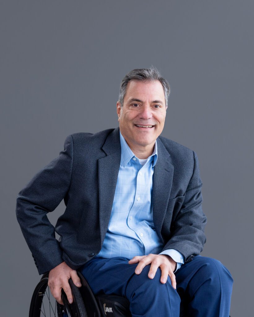 A male public speaker in a wheel chair smiling for his headshot session with a gray backdrop.