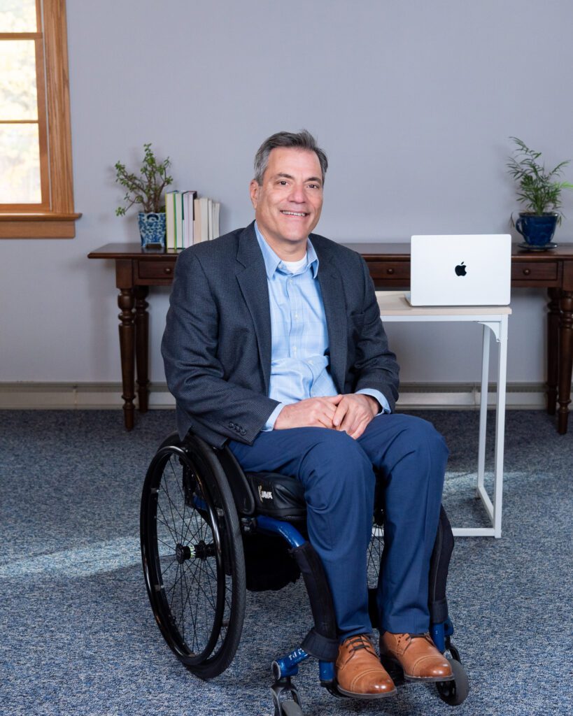 A male public speaker in a wheel chair smiling in front of his desk in his office.
