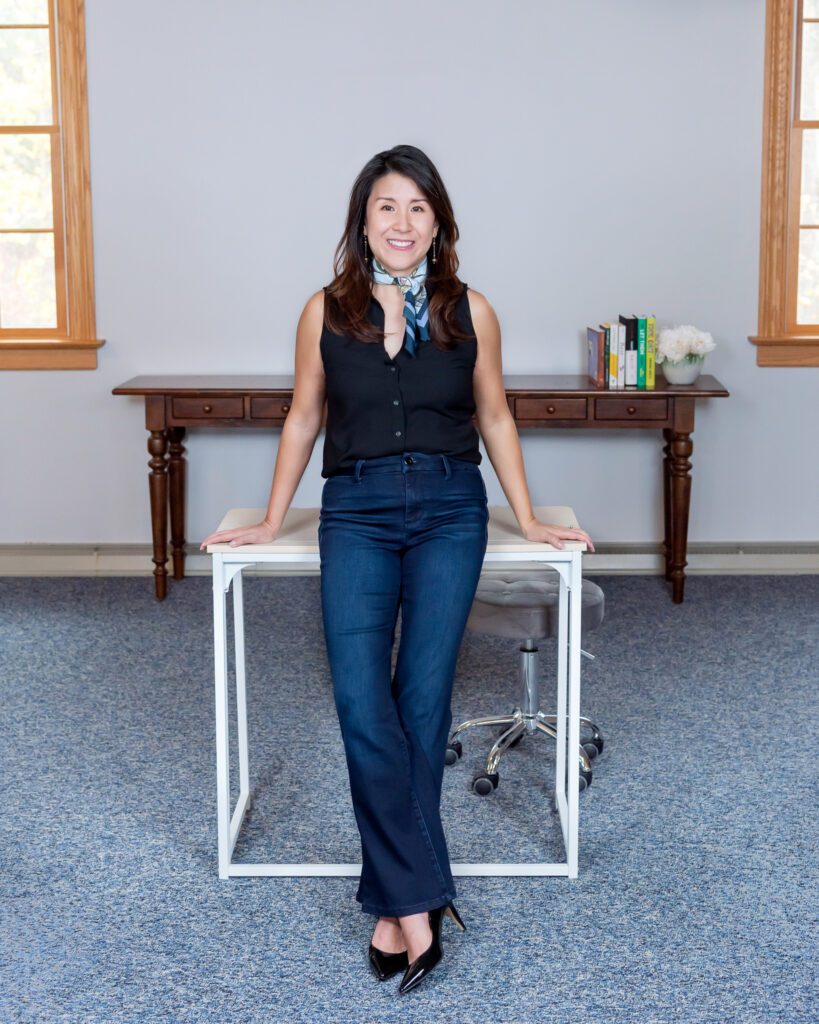 A female Asian executive coach & advisor smiling and leaning on her desk in her office for her indoor lifestyle headshots.
