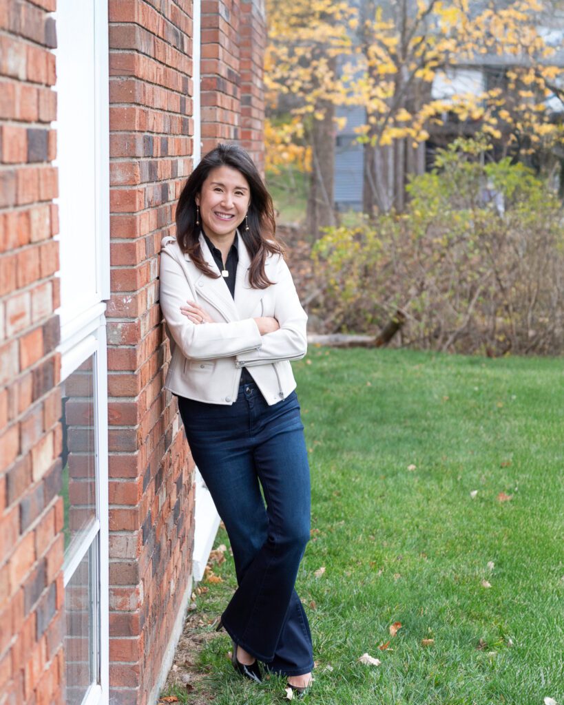 A female Asian executive coach & advisor smiling with her arm crossed, leaning against a brick wall for her outdoor headshot session.