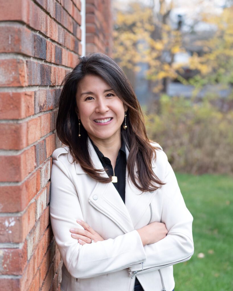 A female Asian executive coach & advisor smiling with her arm crossed, leaning against a brick wall for her outdoor headshot session.