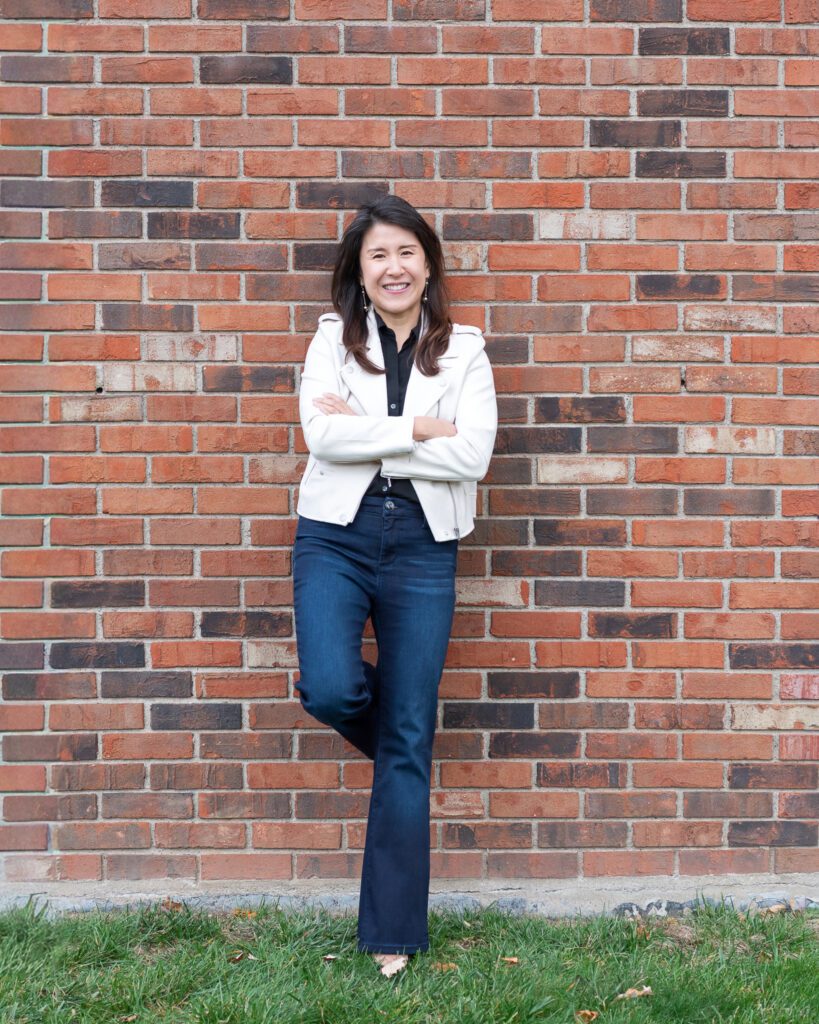 A female Asian executive coach & advisor smiling with her arm crossed, leaning with her back against a brick wall for her outdoor headshot session.