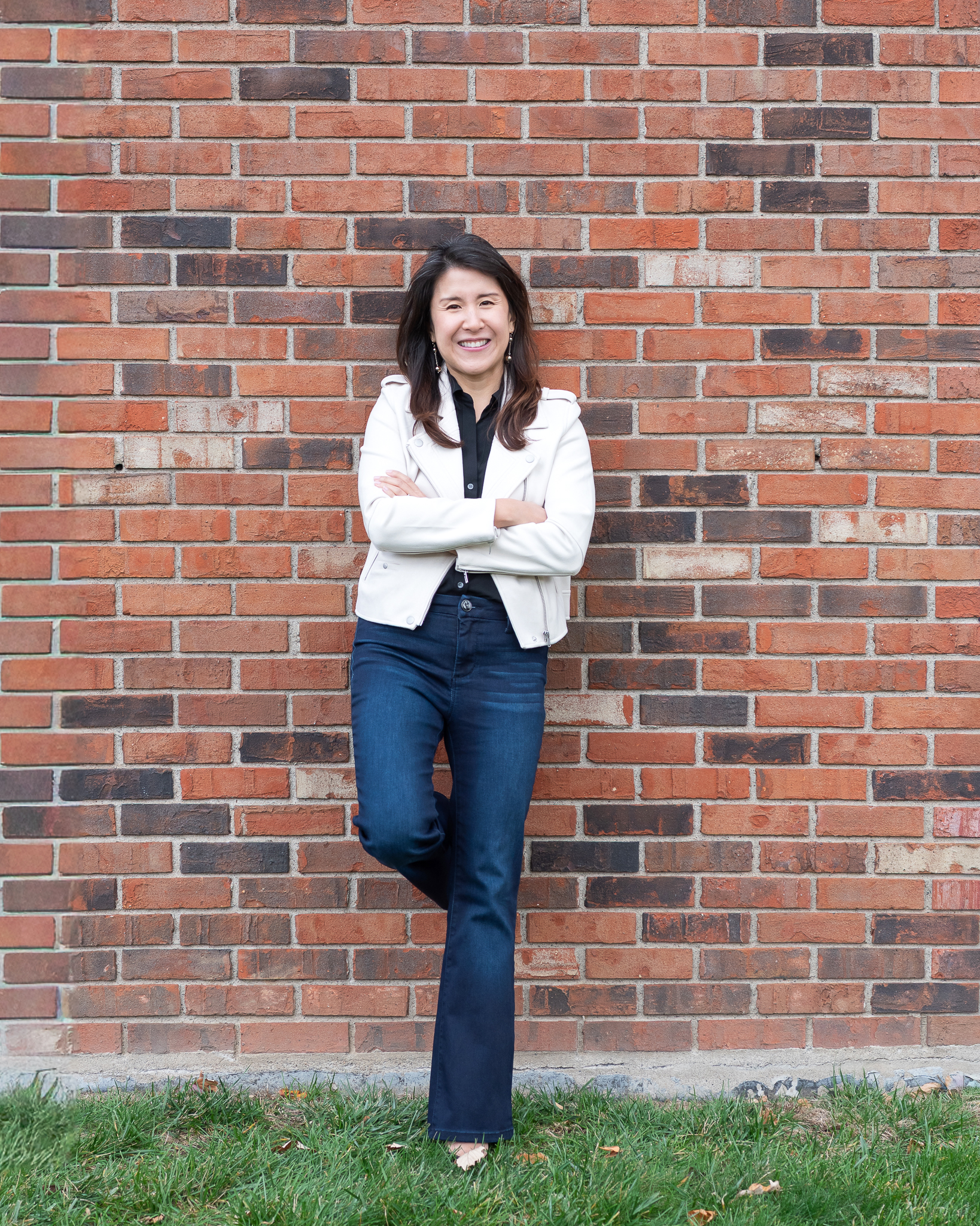A female Asian executive coach & advisor smiling with her arm crossed, leaning with her back against a brick wall for her outdoor headshot session.