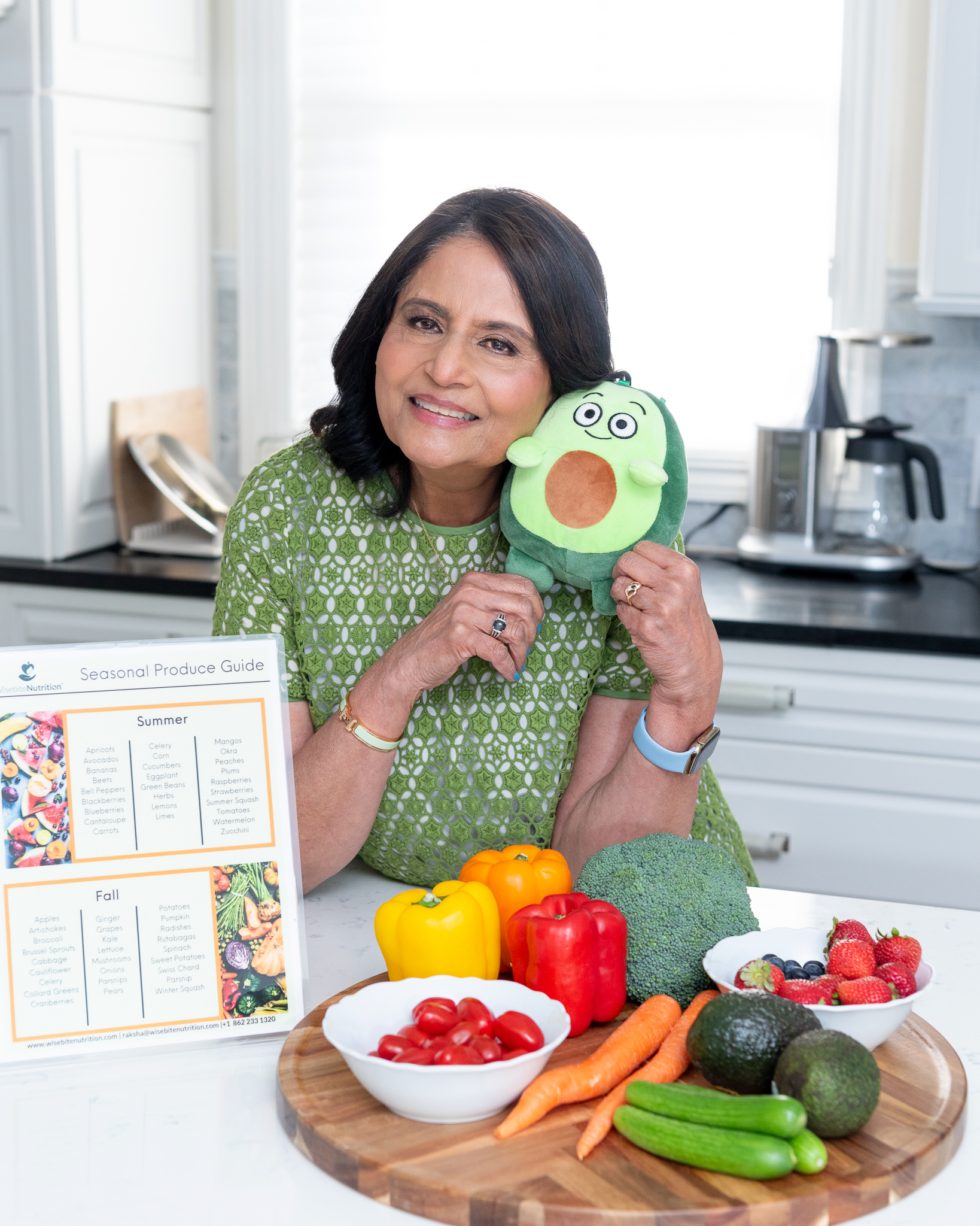 A female nutritionist posing for her branding photoshoot by in front of a platter of fruits and vegetables and holding a cute avocado plushy.
